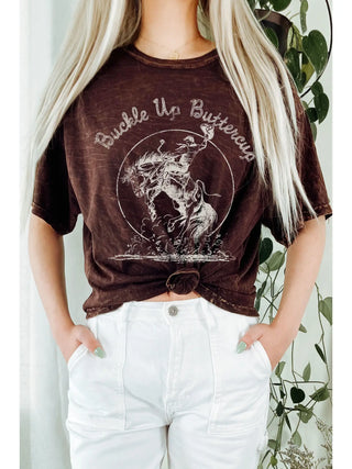 Person wearing a brown graphic t-shirt with text and design, standing against a white background with greenery.