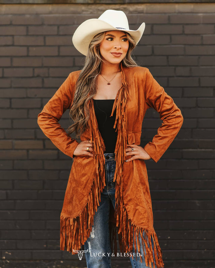 Woman wearing a brown fringe cardigan, black top, jeans, and white cowboy hat against a brick wall.